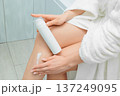Close up hands of woman in a white bathrobe holding a white lotion bottle and applying cream to her legs, performing a daily skincare routine for moisturized and healthy skin 137249095