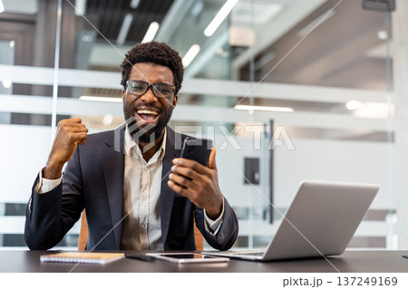 Businessman expressing excitement and confidence while celebrating victory at work, happily receiving good news and positive results on a mobile phone in a modern office setup 137249169