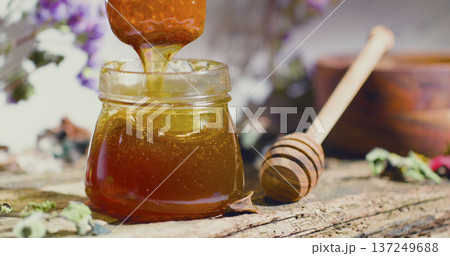 Honey pouring into a jar with a wooden stick on a rustic table surrounded by flowers and natural elements Honey pouring into a jar with a wooden stick on a rustic table surrounded by flowers and natural elements 137249688