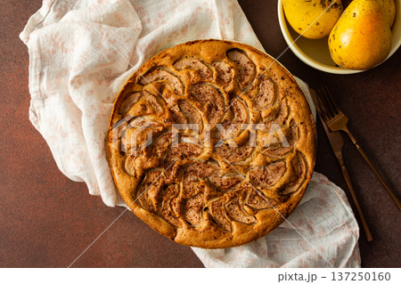 A fresh pear pie is placed on a cloth on a table. Nearby, a bowl holds ripe pears and a small vase has flowers. The setting looks warm and inviting for sharing a treat A fresh pear pie is placed on a cloth on a table. Nearby, a bowl holds ripe pears and a small vase has flowers. The setting looks warm and inviting for sharing a treat 137250160