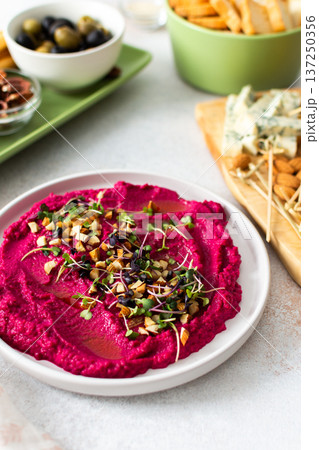 Beetroot hummus is placed in the center of a charcuterie board, surrounded by snacks like olives, nuts, cheese, and crispy breadsticks. A light, inviting setup showcases vibrant colors Beetroot hummus is placed in the center of a charcuterie board, surrounded by snacks like olives, nuts, cheese, and crispy breadsticks. A light, inviting setup showcases vibrant colors 137250356