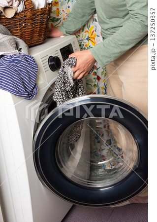 A woman sorts and loads laundry into a washing machine. The vibrant floral wall adds warmth to the cozy room. A basket of clothes sits nearby, ready to be washed 137250357