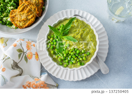 Warm bowls of creamy green pea and broccoli soup are garnished with fresh peas and mint. Crispy zucchini pancakes and a refreshing salad complete this delightful meal 137250405