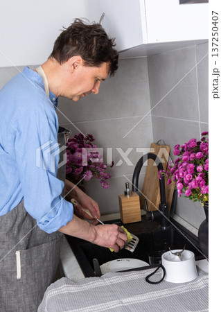 A man stands at a sink in a kitchen, washing dirty dishes and glassware. Flowers sit on the counter as fresh light comes in through the window. The setting is busy and practical 137250407