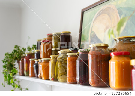 Canned vegetables and fruits are lined up on a shelf in a kitchen, showing winter preparations. A painting of a basket is seen in the background as part of the decor Canned vegetables and fruits are lined up on a shelf in a kitchen, showing winter preparations. A painting of a basket is seen in the background as part of the decor 137250654