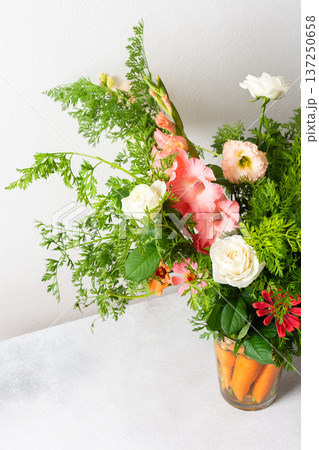 A bouquet of roses, gladioli, zinnias, and eustomas stands in water. Carrots with green tops are part of the arrangement on a plain table against a light background 137250658