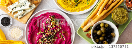 Colorful spread features beetroot, classic, broccoli, and turmeric hummus showcased on a charcuterie board with vibrant toppings, breadsticks, and assorted olives, perfect for gatherings, banner 137250751