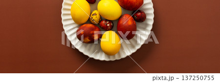 A collection of brightly colored Easter eggs in various sizes and patterns is displayed on a white scalloped plate, resting on a warm brown surface, banner A collection of brightly colored Easter eggs in various sizes and patterns is displayed on a white scalloped plate, resting on a warm brown surface, banner 137250755
