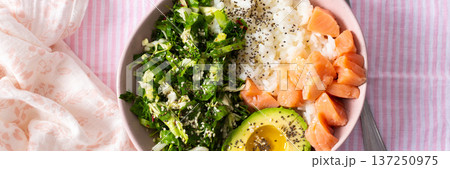 Fresh breakfast bowl featuring salmon, sliced avocado, leafy greens, and rice served on a pink striped tablecloth. Perfect for a nutritious start to the day, banner 137250975