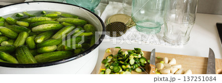 Cucumbers are soaking in a bowl while garlic and cucumber slices are on a cutting board. Several empty jars sit nearby on a clean countertop. It is a preparation activity for winter storage, banner 137251084