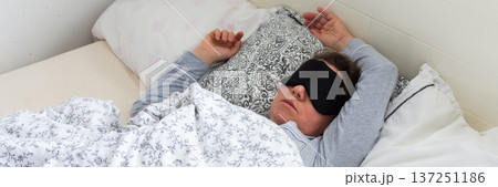A 50-year-old woman is sound asleep in her bed, wearing a sleep mask. She is surrounded by pillows and blankets, creating a tranquil atmosphere. Sunlight gently filters into the room, banner A 50-year-old woman is sound asleep in her bed, wearing a sleep mask. She is surrounded by pillows and blankets, creating a tranquil atmosphere. Sunlight gently filters into the room, banner 137251186
