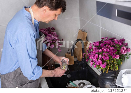 A man stands at a sink in a kitchen, washing dirty dishes and glassware. Flowers sit on the counter as fresh light comes in through the window. The setting is busy and practical A man stands at a sink in a kitchen, washing dirty dishes and glassware. Flowers sit on the counter as fresh light comes in through the window. The setting is busy and practical 137251274