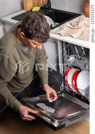 In a kitchen, a man is putting a dishwashing detergent tablet into the dishwasher. He is getting ready to clean the dirty dishes inside the machine In a kitchen, a man is putting a dishwashing detergent tablet into the dishwasher. He is getting ready to clean the dirty dishes inside the machine 137251275