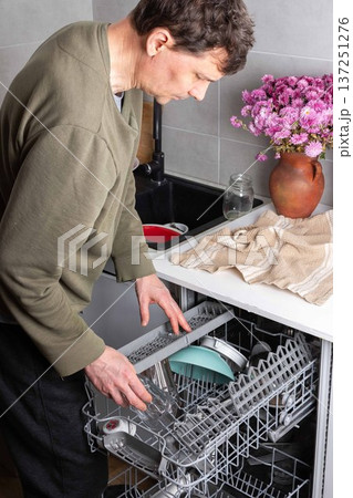 A man is putting dirty dishes into the dishwasher in his kitchen. He focuses on the task while several items rest in the appliance, ready for cleaning 137251276