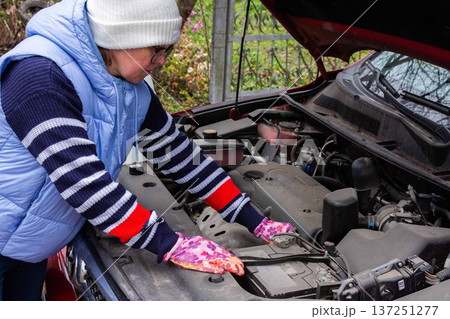 A woman is working on the car engine in a driveway. She is checking the battery and other parts to ensure the vehicle is ready for the trip. It is likely a cool day 137251277