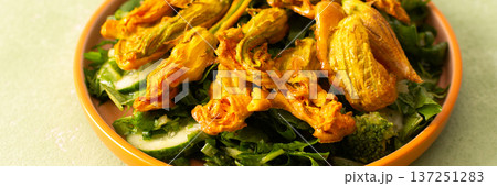 Roasted zucchini flowers are placed on a bed of fresh greens, cucumbers, and broccoli during a summer meal. This dish highlights the flavors of summer ingredients, banner 137251283