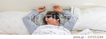 A 50-year-old woman is comfortably sleeping in her bed with a sleep mask on. She is wrapped in a soft blanket, resting peacefully in a serene bedroom setting, banner A 50-year-old woman is comfortably sleeping in her bed with a sleep mask on. She is wrapped in a soft blanket, resting peacefully in a serene bedroom setting, banner 137251284