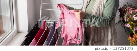 A 50-year-old woman is hanging her laundry on a metal drying rack on her balcony. She looks focused while handling the clothes and has flowers nearby, banner 137251372