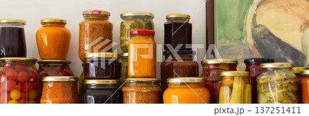 Canned vegetables and fruits are lined up on a shelf in a kitchen, showing winter preparations. A painting of a basket is seen in the background as part of the decor, banner Canned vegetables and fruits are lined up on a shelf in a kitchen, showing winter preparations. A painting of a basket is seen in the background as part of the decor, banner 137251411