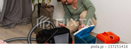 A person is getting ready to vacuum a room. They are attaching a container to the vacuum cleaner while kneeling on the floor. A plant is visible in the background, banner 137251418