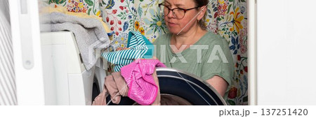 Woman stands in bathroom putting clothes into washing machine while surrounded by towels and laundry, carrying out daily household task with focus and care, banner 137251420