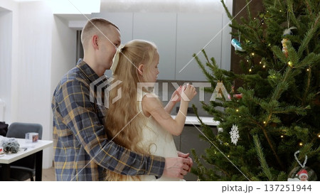 Father embracing his daughter while decorating the christmas tree with ornaments and lights in their modern home, enjoying a warm family moment during the festive season Father embracing his daughter while decorating the christmas tree with ornaments and lights in their modern home, enjoying a warm family moment during the festive season 137251944