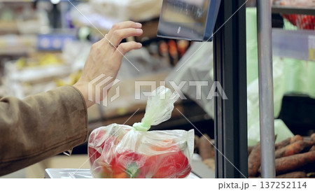 Customer using digital scale to weigh fresh produce in plastic bag at grocery store, choosing nutritious ingredients for cooking Customer using digital scale to weigh fresh produce in plastic bag at grocery store, choosing nutritious ingredients for cooking 137252114