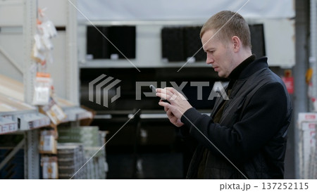 A man in a hardware store, choosing flooring. Shopper using a smartphone to compare prices, search for products, and read reviews while browsing in a home improvement store aisle A man in a hardware store, choosing flooring. Shopper using a smartphone to compare prices, search for products, and read reviews while browsing in a home improvement store aisle 137252115
