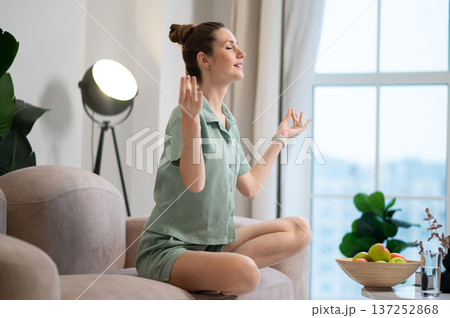 Young woman sitting on the sofa in lotus pose and meditating Young woman sitting on the sofa in lotus pose and meditating 137252868