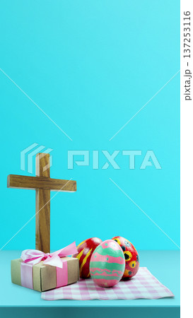A wooden cross stands next to three painted Easter eggs and a small gift box tied with a pink ribbon. The scene is set on a light blue background with a pink checkered napkin A wooden cross stands next to three painted Easter eggs and a small gift box tied with a pink ribbon. The scene is set on a light blue background with a pink checkered napkin 137253116