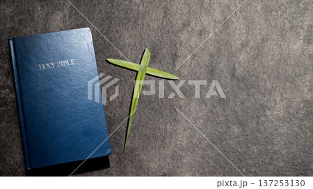A blue Holy Bible and a handmade palm leaf cross on a dark textured stone background. A symbolic composition for Palm Sunday, Holy Week, and Christian faith celebrations with copy space 137253130