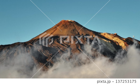 Tenerife, Canary Islands: Low clouds covering Mount Teide's slopes create a breathtaking sunrise scene. Towering peak of a volcano against clear blue sky, stunning landscape. Static shot 137253175