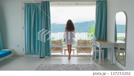 Woman in white tshirt walking hotel room balcony window, enjoy lake surrounded by lush green mountains and cloudy sky, enjoying a peaceful morning view during her tropical vacation to Bali. Back view 137253202