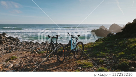 Portugal, Lisbon: Two bicycles parked on a rocky trail overlooking a vibrant ocean coast beach with crashing waves and distant cliffs, representing adventure, travel, and environmental exploration. 137253210