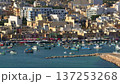 Malta, Marsaxlokk Harbour: Colorful Luzzu boats floating in blue water, with market stalls and residential buildings houses creating a vibrant scene. Travel, tourism, vacation. Aerial drone shot Malta, Marsaxlokk Harbour: Colorful Luzzu boats floating in blue water, with market stalls and residential buildings houses creating a vibrant scene. Travel, tourism, vacation. Aerial drone shot 137253268