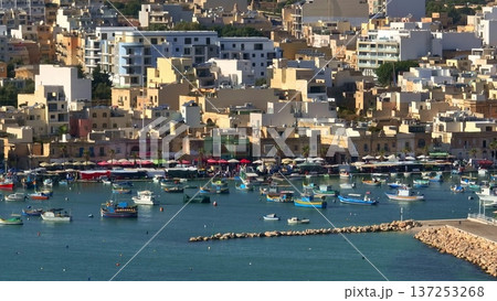 Malta, Marsaxlokk Harbour: Colorful Luzzu boats floating in blue water, with market stalls and residential buildings houses creating a vibrant scene. Travel, tourism, vacation. Aerial drone shot 137253268