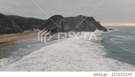 Portugal, Lisbon: Praia da Adraga beach presenting beautiful rugged cliffs meeting the powerful Atlantic ocean, with foamy waves rolling onto the sand on a cloudy dramatic day. Aerial drone flight Portugal, Lisbon: Praia da Adraga beach presenting beautiful rugged cliffs meeting the powerful Atlantic ocean, with foamy waves rolling onto the sand on a cloudy dramatic day. Aerial drone flight 137253279