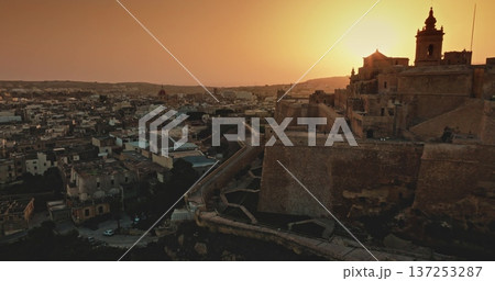 Malta, Comino island: Warm sunset light illuminating the Cittadella bastions in Gozo, Malta, blending history with a modern urban landscape. Drone flight over evening city Malta, Comino island: Warm sunset light illuminating the Cittadella bastions in Gozo, Malta, blending history with a modern urban landscape. Drone flight over evening city 137253287