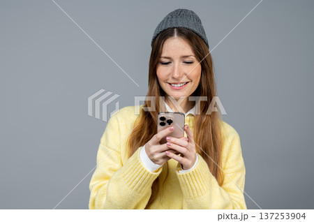 Smiling and contented young woman in grey hat texting on her phone and feeling good Smiling and contented young woman in grey hat texting on her phone and feeling good 137253904