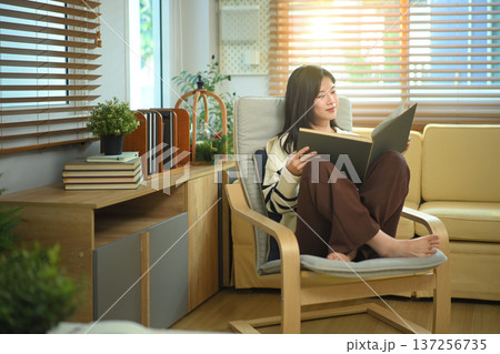 Happy relaxed young woman resting on armchair reading book and holding coffee cup 137256735