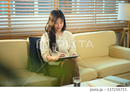 Woman using digital tablet with stylus pen on sofa in living room Woman using digital tablet with stylus pen on sofa in living room 137256755