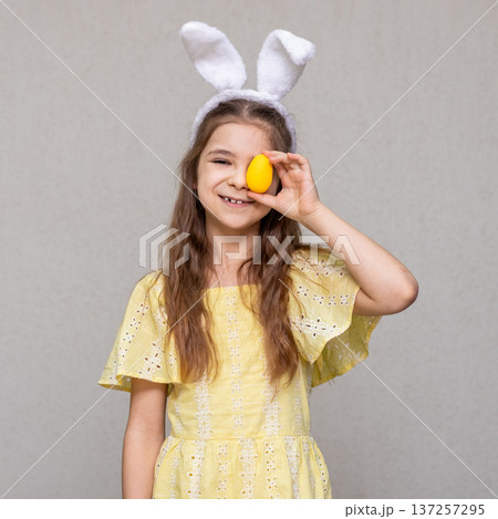 Smiling girl in bunny ears holding a yellow Easter egg and wearing a yellow dress 137257295
