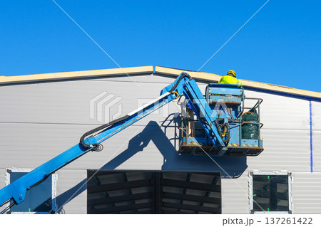 Construction Worker on Hydraulic Lift Installing Roof Panels on Industrial Building Construction Worker on Hydraulic Lift Installing Roof Panels on Industrial Building 137261422