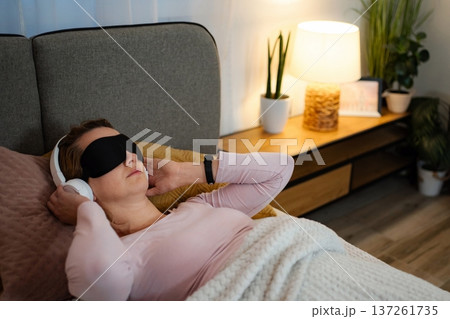 Woman relaxing in bed listening to music with sleep mask Woman relaxing in bed listening to music with sleep mask 137261735