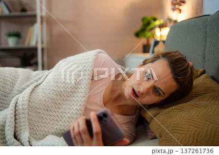 Woman feeling shocked reading shocking news on smartphone in bed Woman feeling shocked reading shocking news on smartphone in bed 137261736