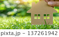 Hand holding a house cutout over lush green grass in a bright, sunlit garden setting 137261942