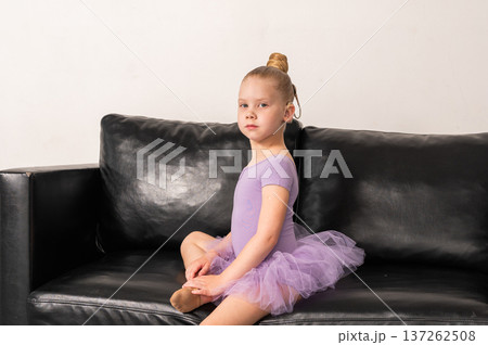Young ballerina with a cochlear implant sits on a black couch during a quiet rehearsal break. Inclusive ballet, hearing technology, and focused childhood through rest and reflection. Young ballerina with a cochlear implant sits on a black couch during a quiet rehearsal break. Inclusive ballet, hearing technology, and focused childhood through rest and reflection. 137262508