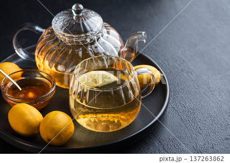 Glass teapot with amber tea, teacup with lemon slice, and bowl of honey with golden spoon on dark concrete background Glass teapot with amber tea, teacup with lemon slice, and bowl of honey with golden spoon on dark concrete background 137263862