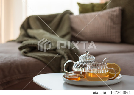 Glass teapot with amber tea, teacup with lemon slice, and bowl of honey with golden spoon on white table in cozy living room Glass teapot with amber tea, teacup with lemon slice, and bowl of honey with golden spoon on white table in cozy living room 137263866