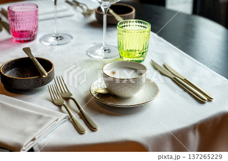 Cappuccino on white linen table, gold cutlery and colored glasses, textured porcelain cup with creamy foam, 137265229